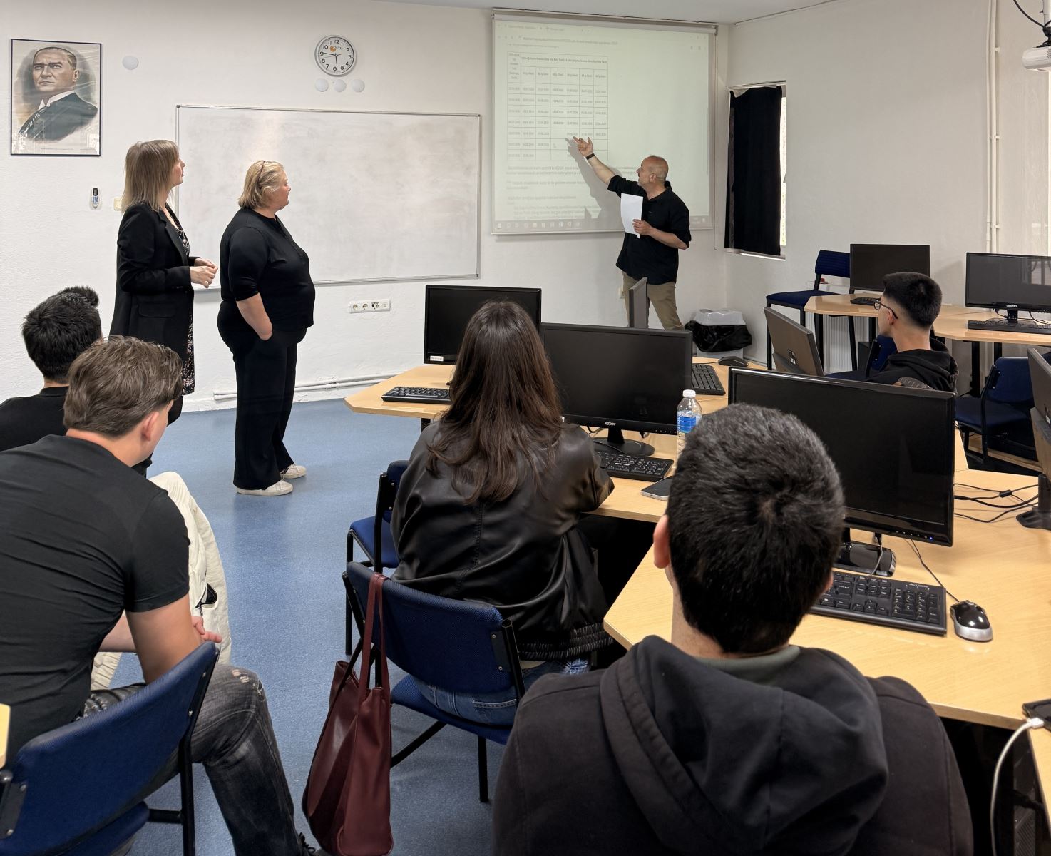 İki kadın bir erkek akademik personel Atatürk portresi önünde ve tahtada karşılarındaki 2025-2026 stajı hakkında öğrencilere bilgi veriyor. 