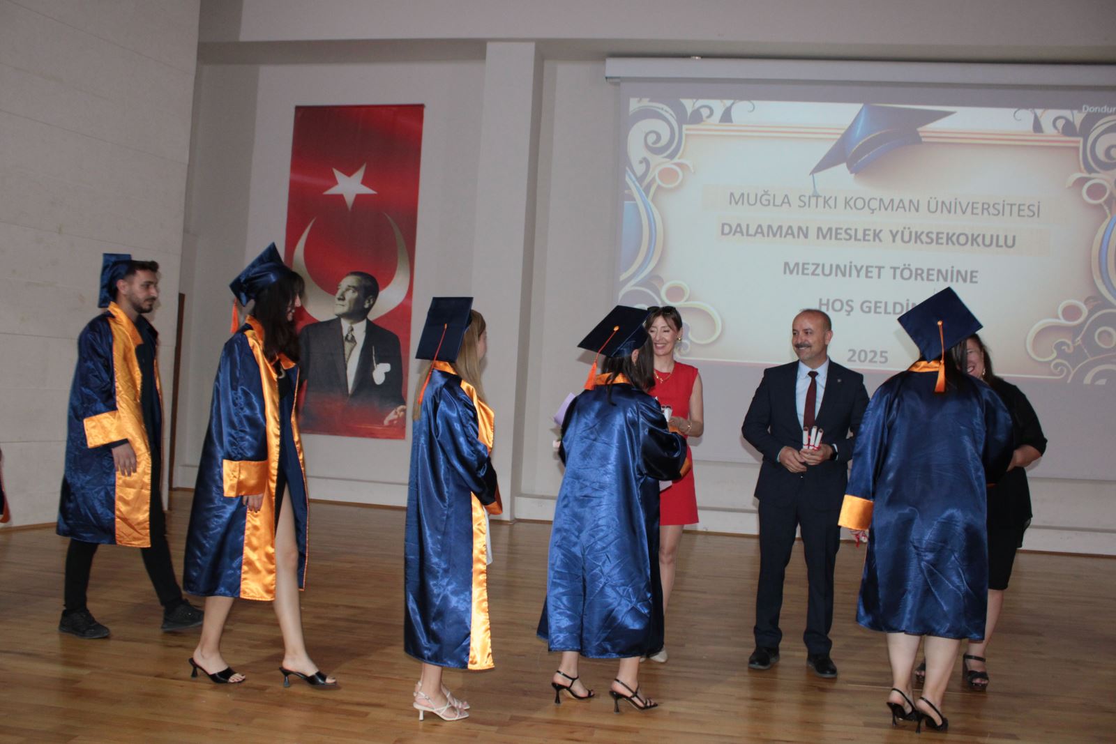 3 öğretim elemanı yüzleri objektife dönük halde, önlerinden geçen öğrencilere başarı belgelerini verip tebrik ediyor. 