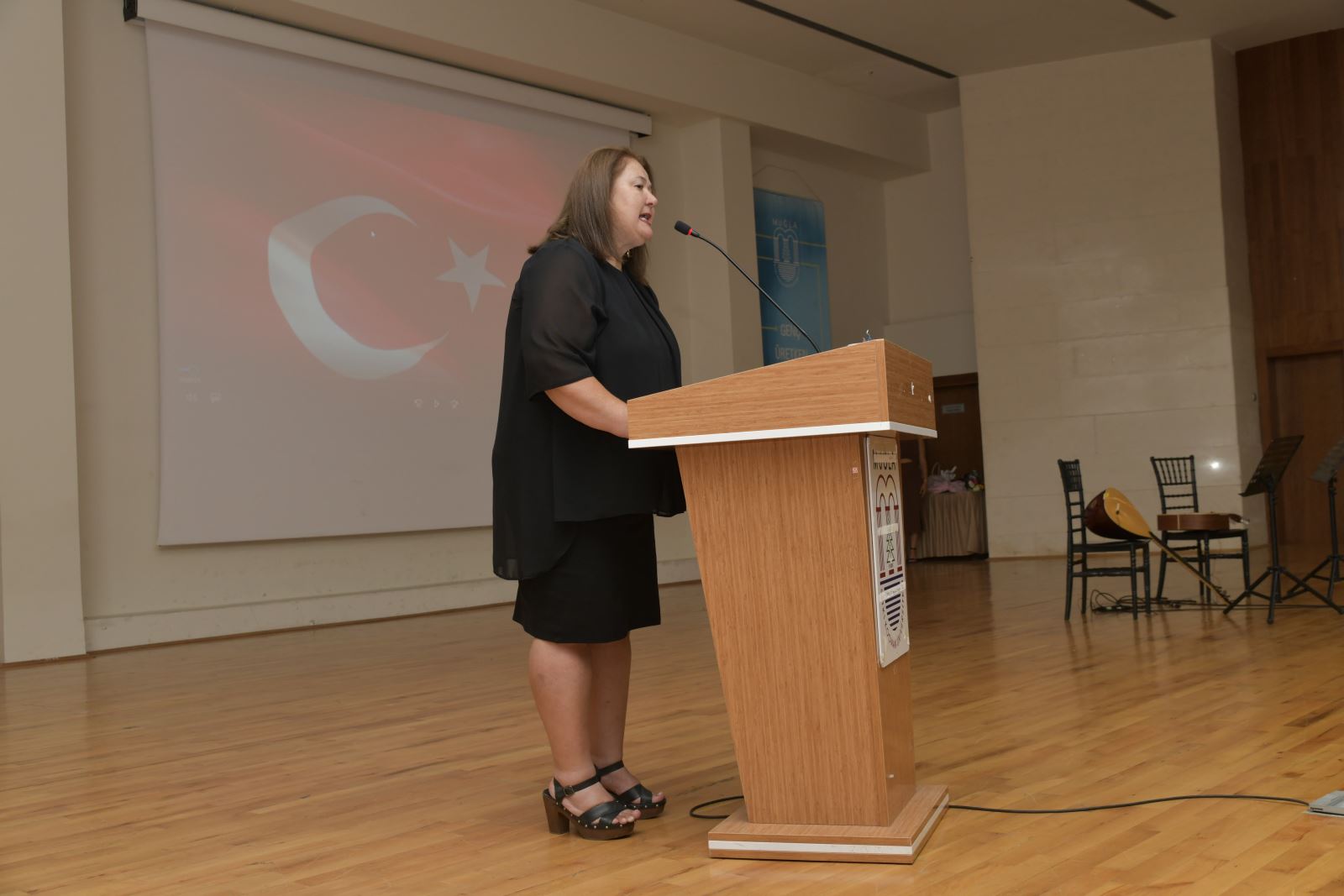 Arkada Türk Bayrağı bulunan bir salonda siyah elbise giymiş bir kadın sunucu konuşuyor