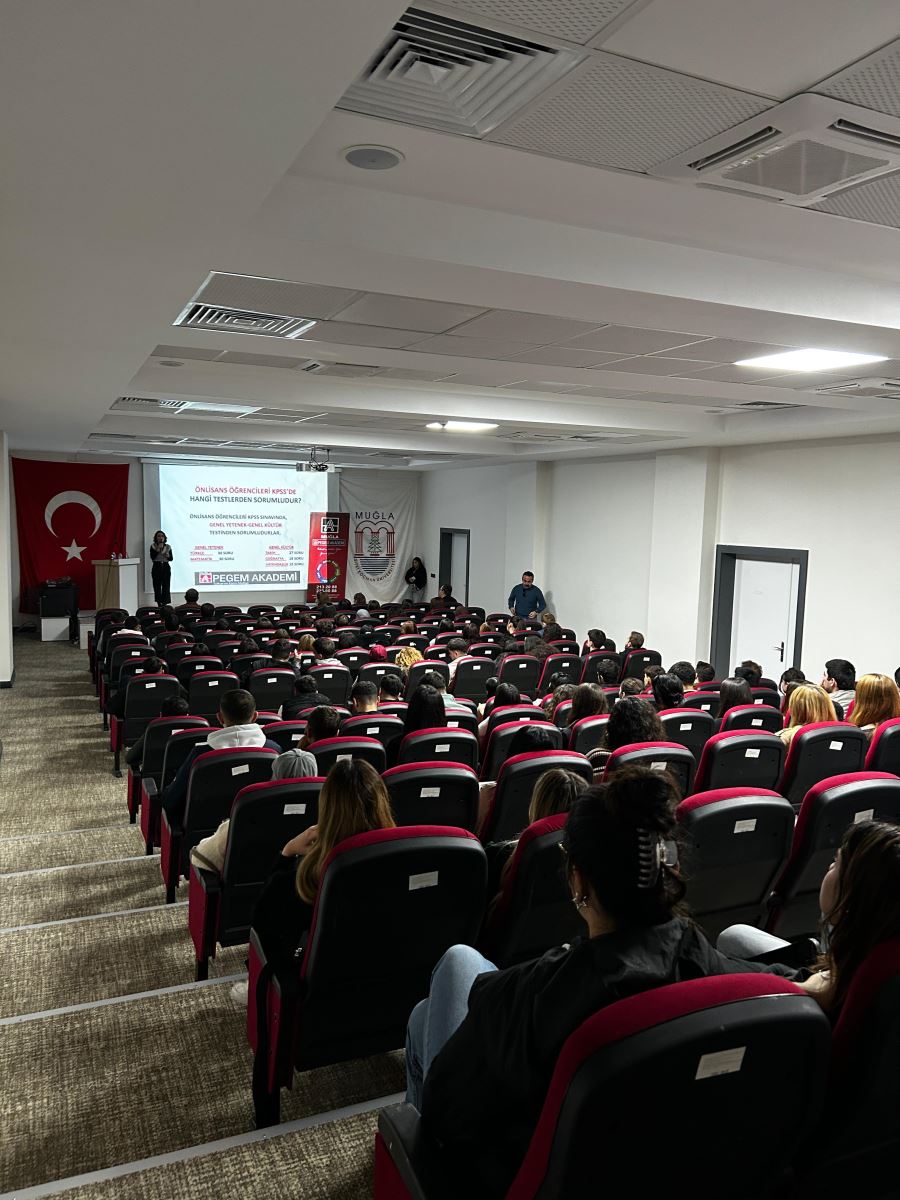 Bu fotoğraf yine salonun arka tarafından çekilmiş ve sahnede başka bir sunum slaydı görünüyor. Slaytta "Önlisans Öğrencileri KPSS'de Hangi Testlerden Sorumludur?" başlığı yer alıyor. Öğrenciler sıralar halinde oturmuş ve sunuma dikkat kesilmiş durumda. Salon düzenli, aydınlatma parlak ve sahnede bir konuşmacı ile yanında bir görevli bulunuyor. Türkiye bayrağı ve Muğla afişi yine sahnenin arkasında yer almakta.