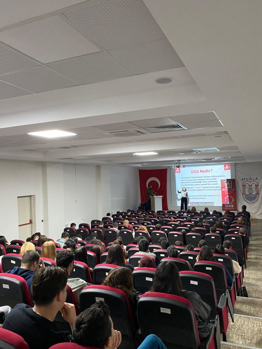 Bu fotoğrafta sahnede bir konuşmacı sunum yapıyor. Sunum ekranında “DGS Nedir?” başlıklı bir slayt görünüyor. Öğrenciler salonun tamamına yayılmış şekilde arkadan öne doğru oturuyor. Sahnenin sol tarafında büyük bir Türk bayrağı, sağ tarafında ise Muğla ile ilgili bir afiş bulunuyor. Tüm koltuklar dolu ve öğrenciler dikkatle sunumu izliyor.